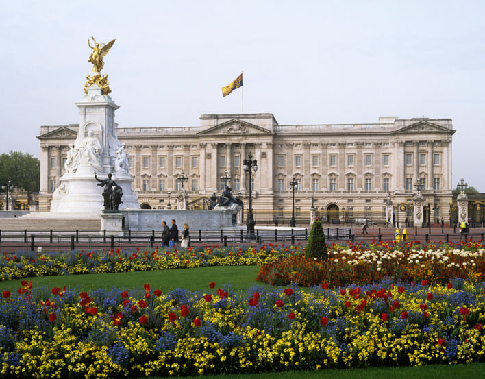 The grey stone facade of Buckingham Palace with gates and flower beds