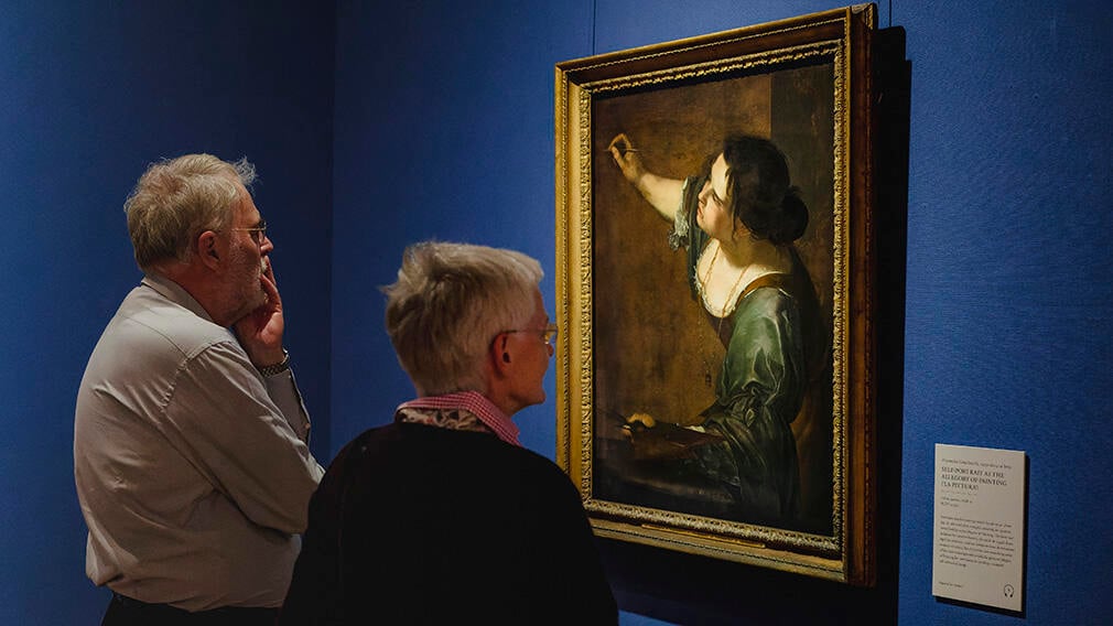 Visitors looking at Artemisia Gentileschi's Self-Portrait during an exhibition (RCIN 405551). A man and a woman looking at a painting hanging on the wall. They both have grey hair.