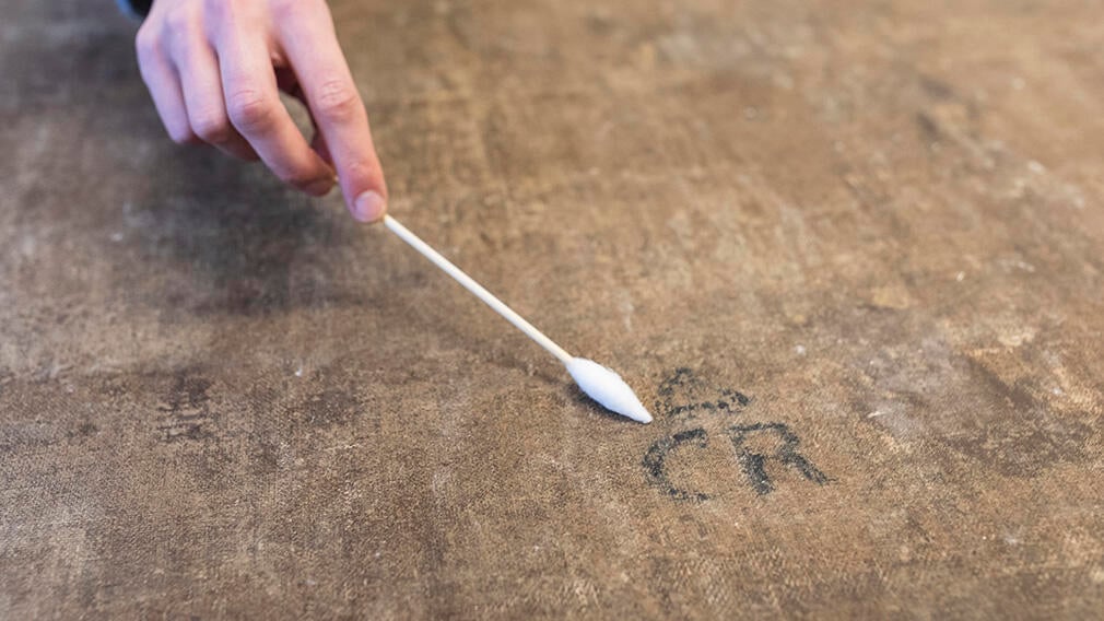 The ‘CR’ brand revealed during conservation treatment. A person holding a cotton bud cleans the surface of the back of a canvas.