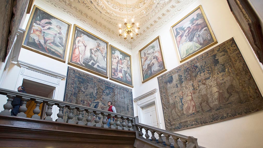 The Great Stair at the Palace of Holyroodhouse A man walks along a landing of a stone staircase. The walls are filled with tapestries and paintings.