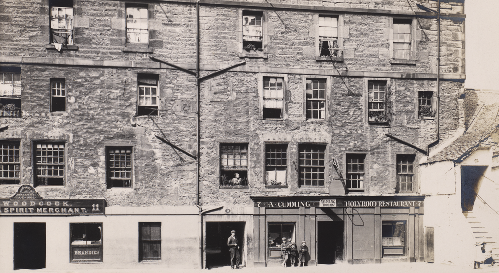 By 1903 the Abbey Strand buildings were very overcrowded, housing up to 25 families. A black and white photo of building in 1903. A man and children stand outside the buildings.